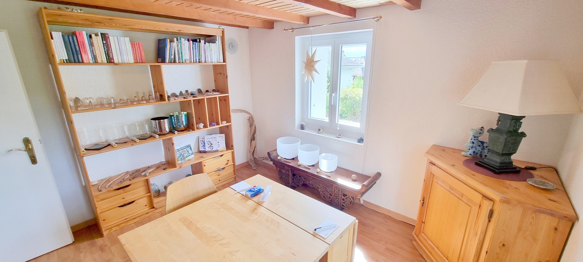 table au centre, étagère avec des livres et des bols sur la gauche, fenêtre au centre, meuble bas avec bols en cristal sous la fenêtre, meuble d&apos;angle à droite avec une lampe à pied